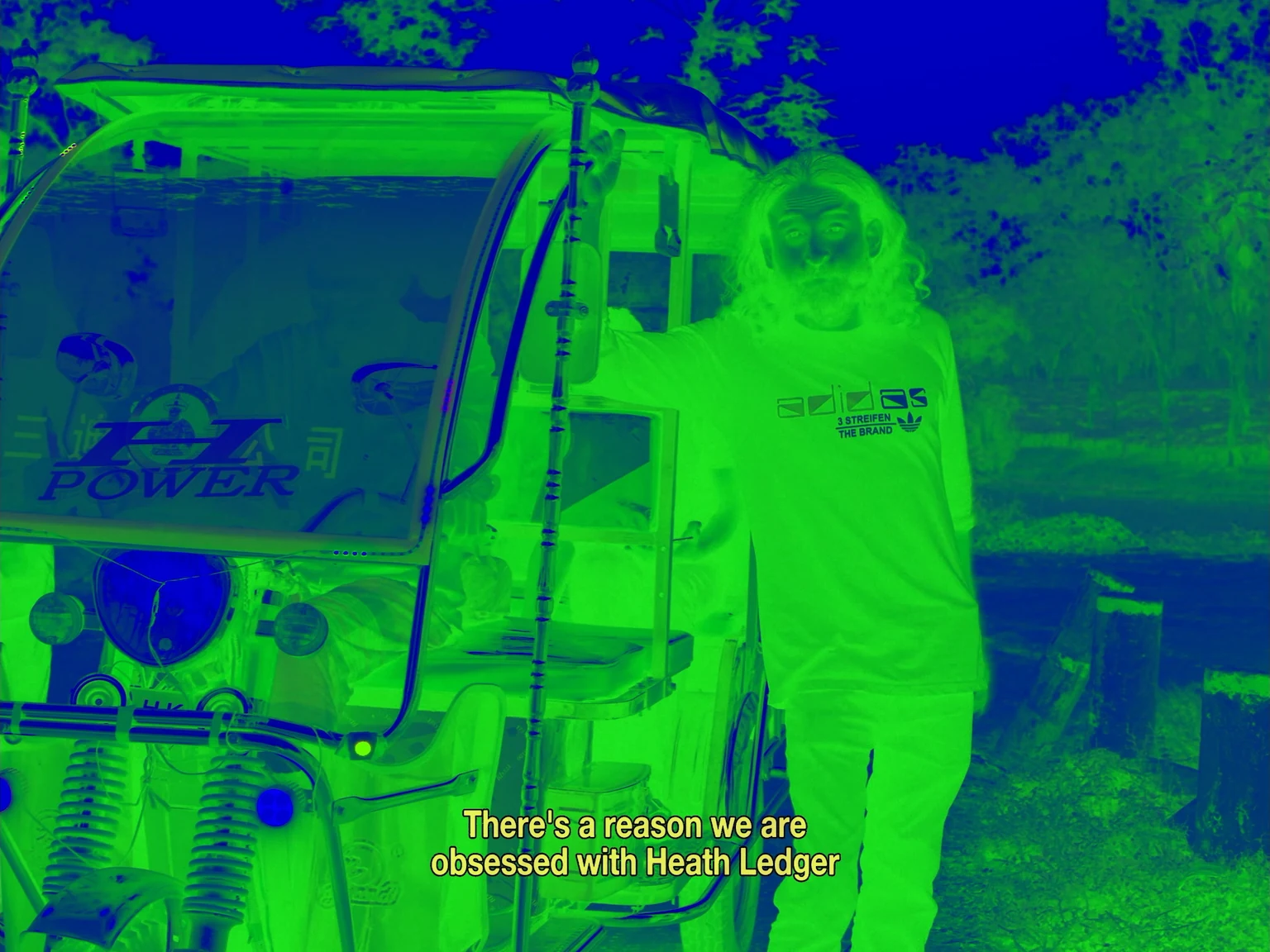 A bearded bandit beside a decorated three-wheeler in an inverted green-and-blue film still