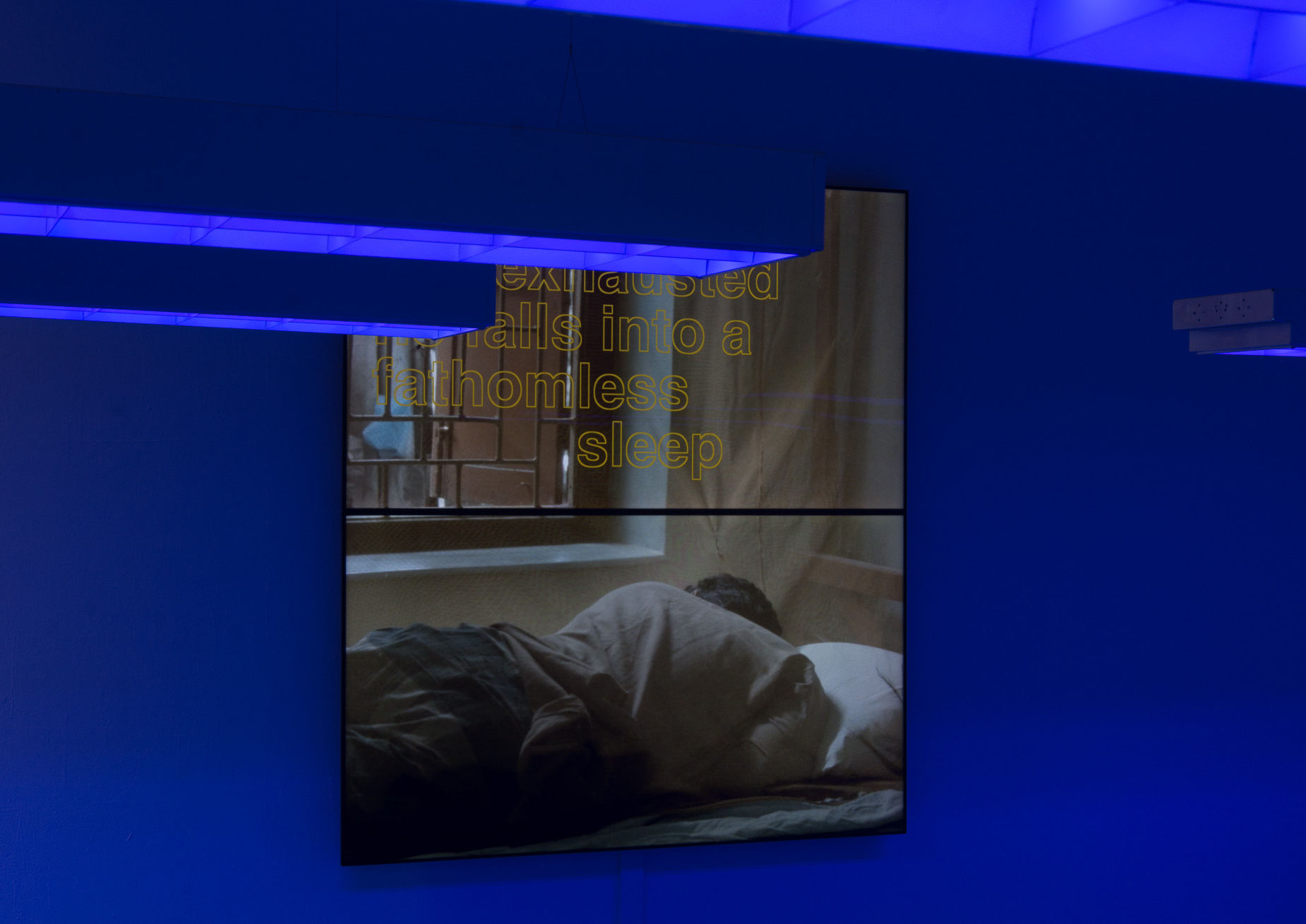 Blue-lit installation with suspended light fixtures and a film projection